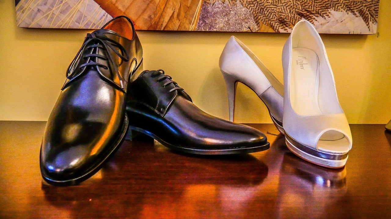 Black leather derby shoes seen from the front and side, with a beige peep-toe heel beside them